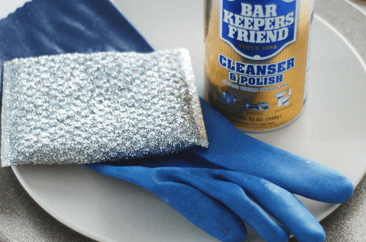 A canister of Bar Keepers Friend cleanser and polish on top of a plate with a dish glove and steel wool sponge