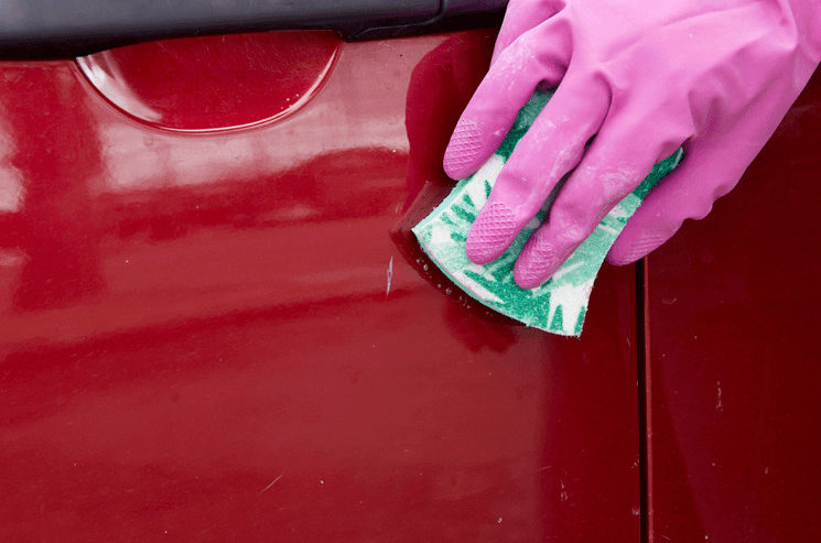 A gloved hand using the soft side of a sponge and Bar Keepers Friend to fix small scratches on a red car door