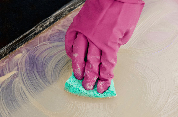 A gloved hand cleaning an oven door window using a sponge and Bar Keepers Friend