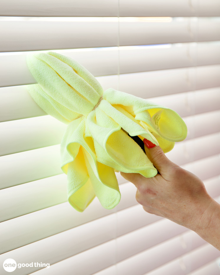 woman's hand using kitchen tongs and a yellow microfiber cloth to dust mini blinds