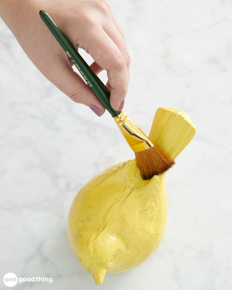 woman's hand using an artist's paint brush to dust a small yellow ceramic bird figure