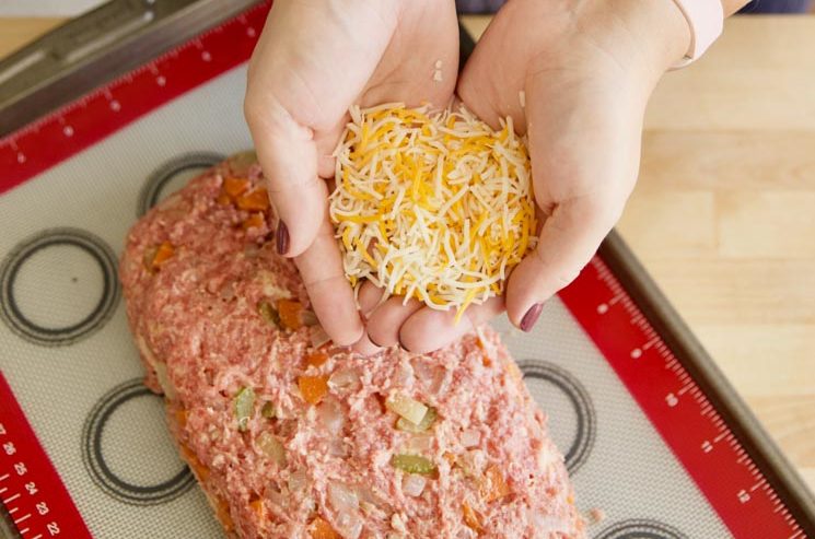 hands holding shredded cheese above raw meatloaf